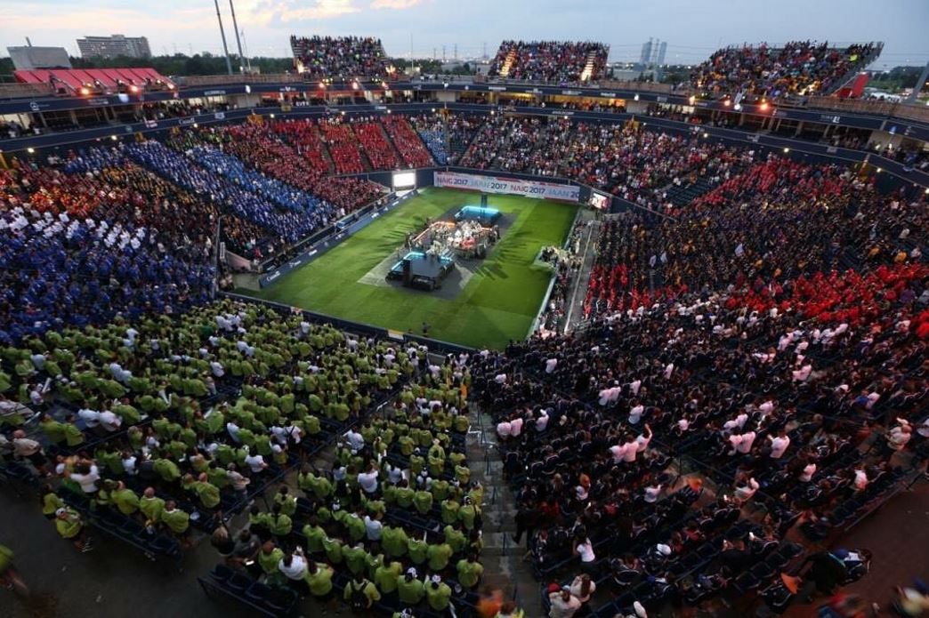 Wide angle photo of an open-air stadium at dusk, filled with spectators watching the opening ceremonies for the North American Indigenous Games on a green field.