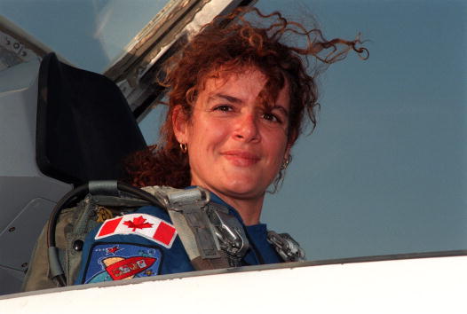 Head-shot of Julie Payette sitting in the open cockpit of a plane. She is wearing a blue uniform and parachute vest with a Canadian flag patched on the shoulder.