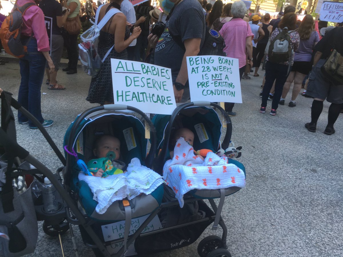 A photo of two babies in two separate strollers. Behind them are protest signs reading "All babies deserve healthcare" and "being born at 28 weeks is not a pre-existing condition."