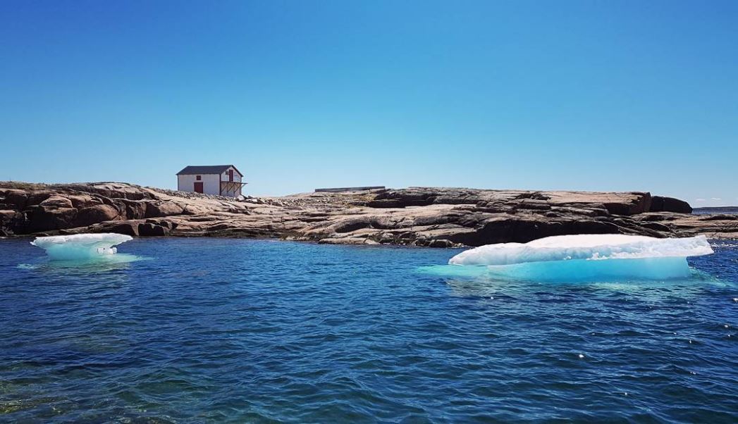 Wide angle shot of a small cabin on a rocky surface under a blue sky. Dark blue water lapping on shore in the foreground with two small icebergs floating nearby.