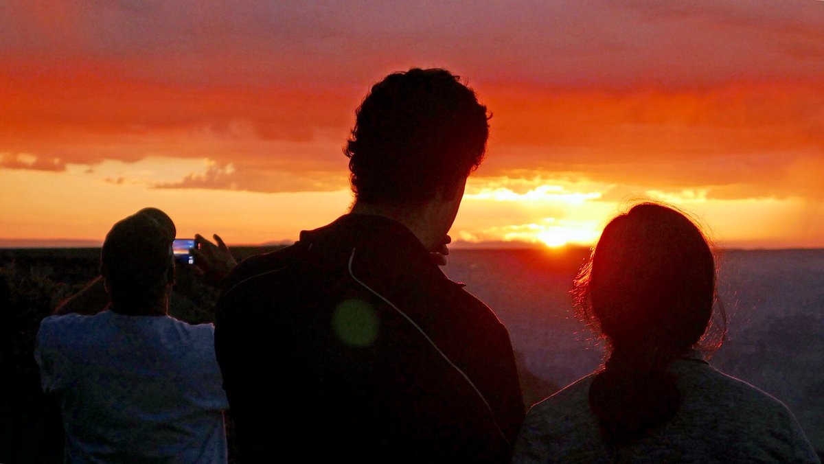 Three people watching sunset as the sun is on the horizon and the clouds are a reddish orange color