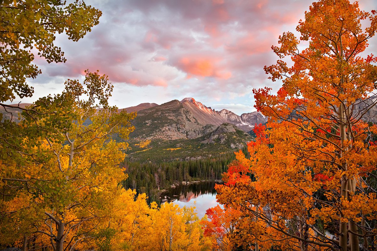 Looking through a gap in fall colored trees is a mountain peak with a lake at its base.