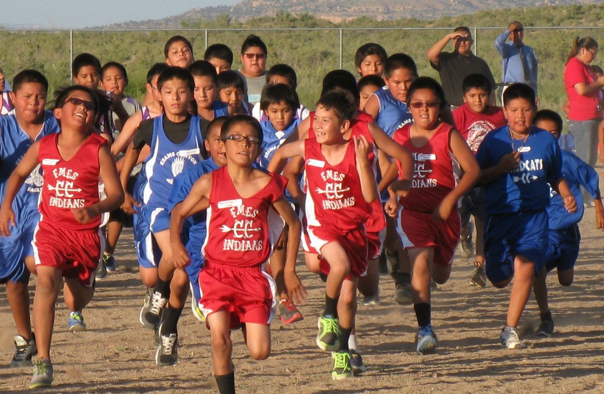 A group of boys in red and blue athletic clothing runs across a dirt playing field.