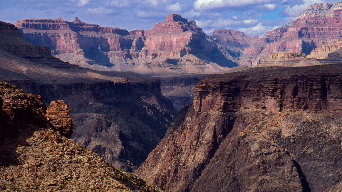 a flat plateau on the horizon drops with sloping sides down into a river gorge. Above the horizon, distant peaks and mesas rise up towards the blue sky.] NPS/Tom Bean.