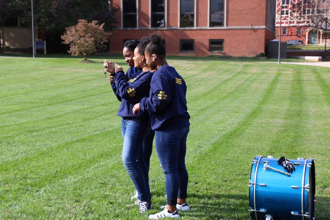 [Image description: Three Bison Song members stand next to each other as they look at the photo showed on their iPhone after the photo was taken.]