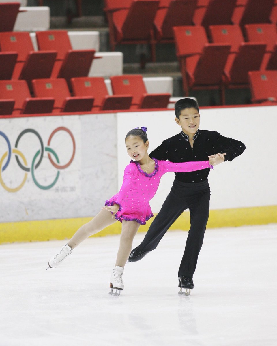 In 2004, tiny little Maia and Alex compete at the Lake Placid Ice Dance Championships. Maia wears a hot pink dress and Alex wears a black studded shirt. They both grin widely at the camera with the Olympic rings displayed on the wall behind them. Photo courtesy of Alex Shibutani.