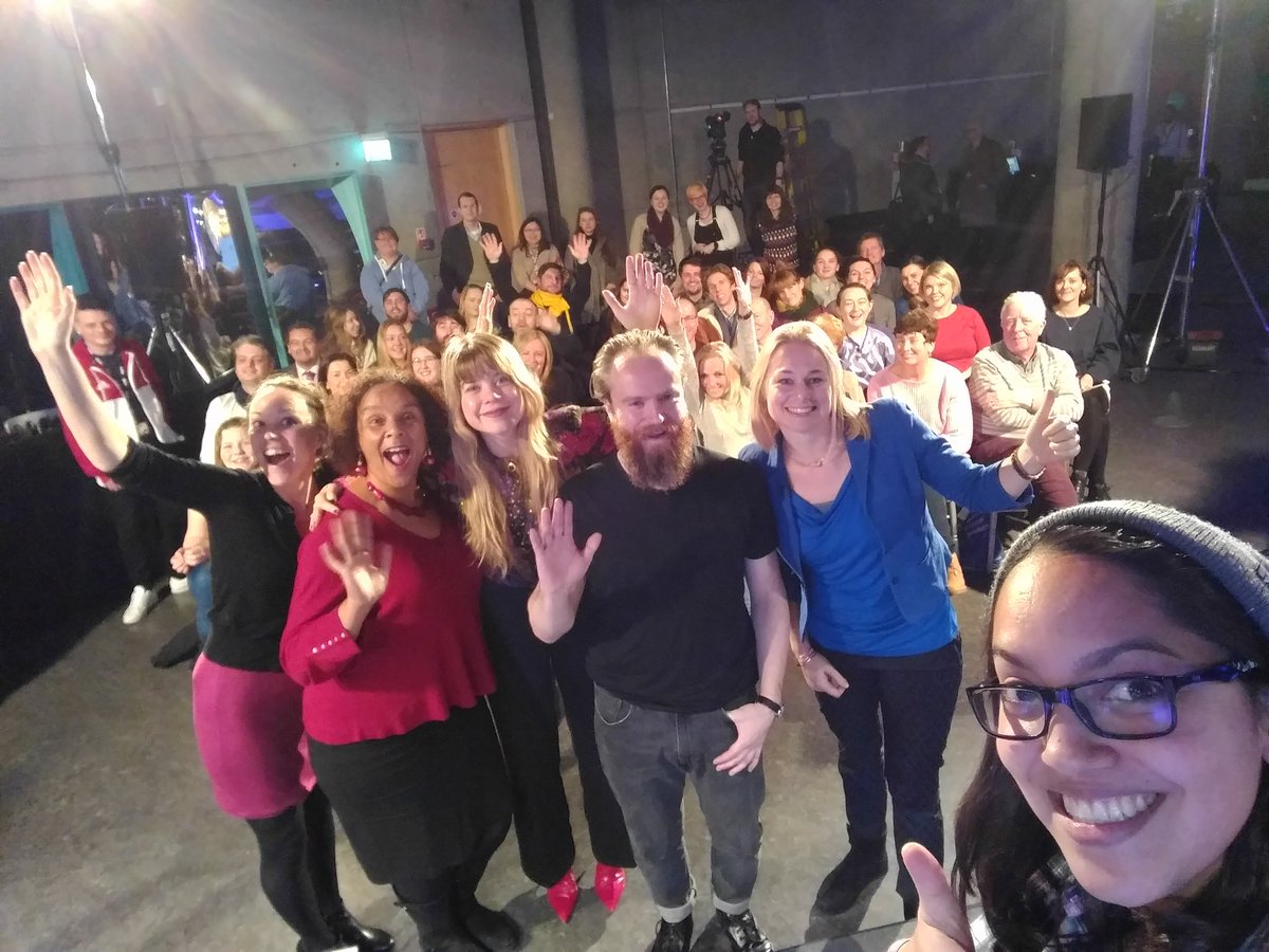 a selfie photo taken from a stage with the photographer in the bottom right corner, 5 people standing in a row, and an audience of people seated behind them.