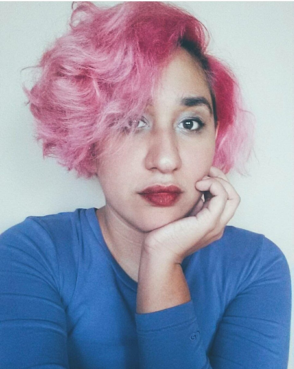 chest up portrait of Annie, she has short, wavy, pink hair, is wearing eye make up and red lip stick. She is wearing an indigo long sleeve shirt and is resting her chin on her left hand and has a blank expression]