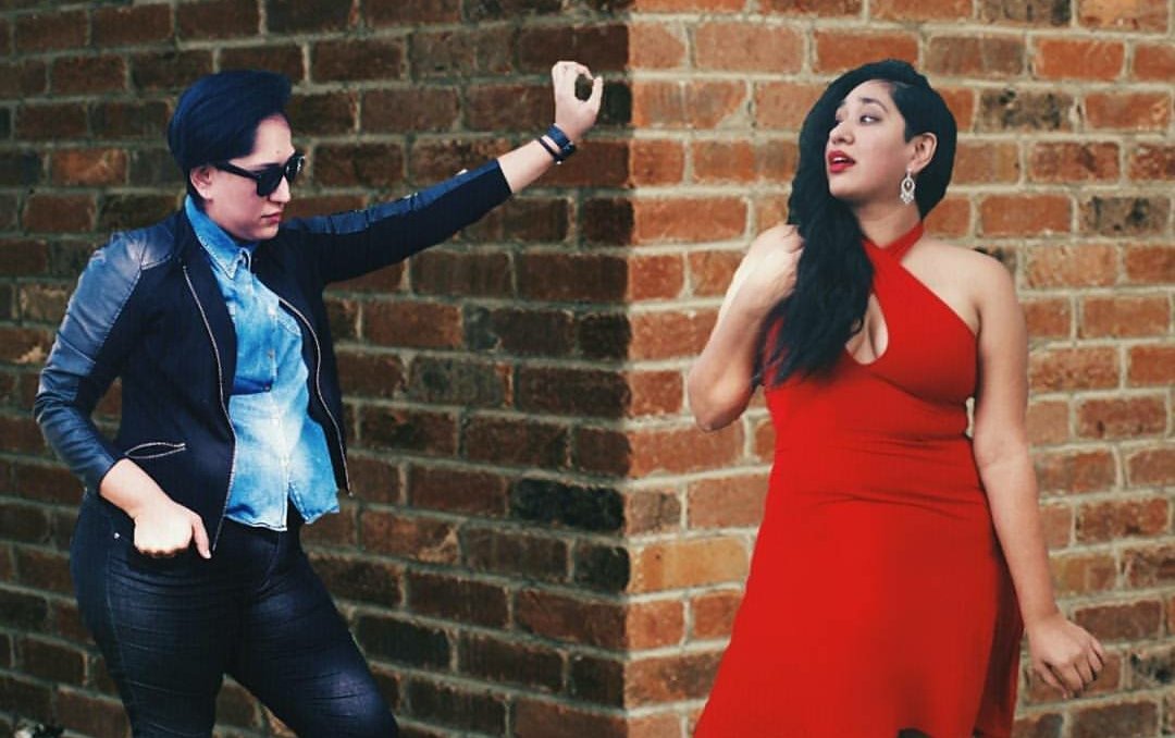 an altered photo, outdoors at the corner of a red brick wall. On one side and in shadow is Annie with short black hair, sunglasses, a denim button-up and black jeans and jacket, she is leaning against the wall. On the other side of the wall and in sunlight is a duplicate version of Annie with long black hair and a form fitting red dress. She is holding her thick hair over to one side
