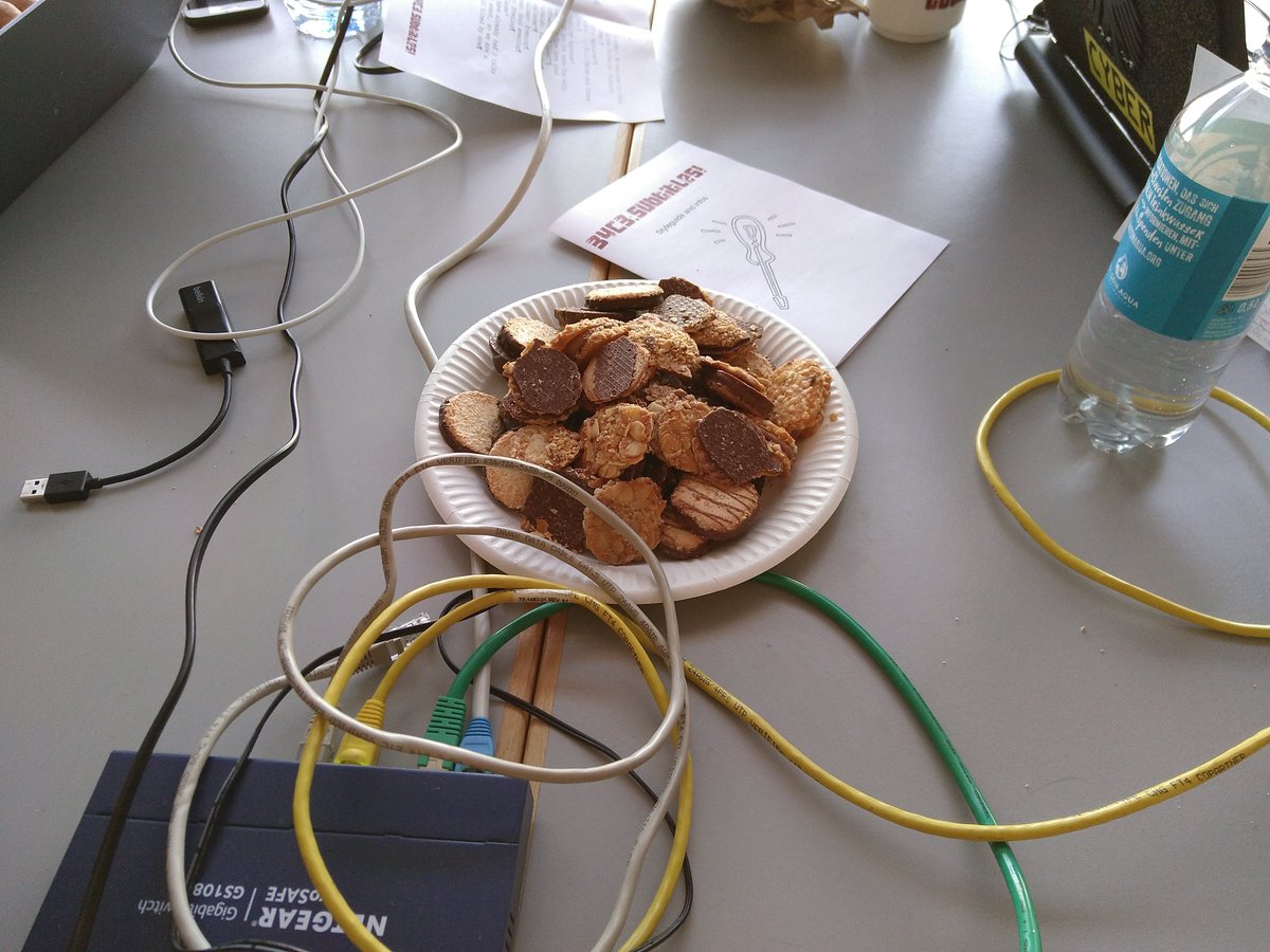 A plate with lots of cookies amongst laptops, a network switch, subtitling style guides, and a bottle of water