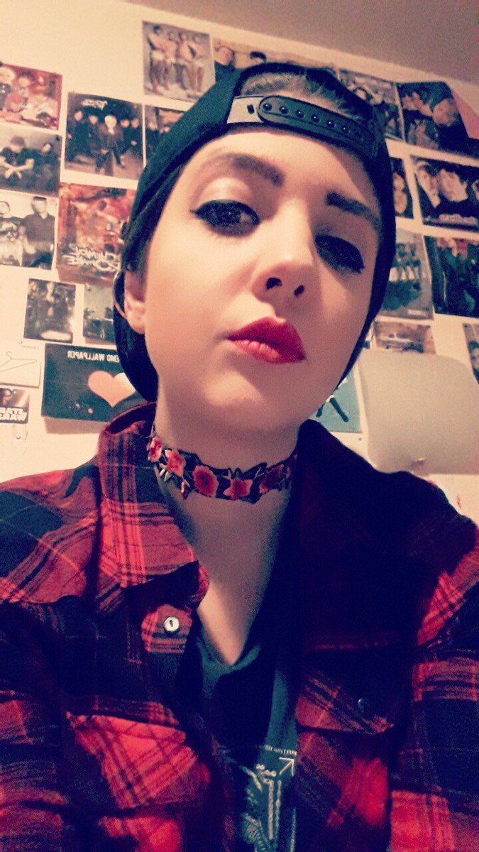 portait picture of me, a young white woman, in front of a wall with emo band posters, wearing a red plaid shirt, a flowery choker necklace, and a black snapback hat front-to-back, with red lipstck and strong eyeliner