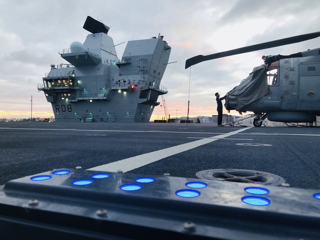 Two silhouetted figures remove the covers from a Merlin helicopter.