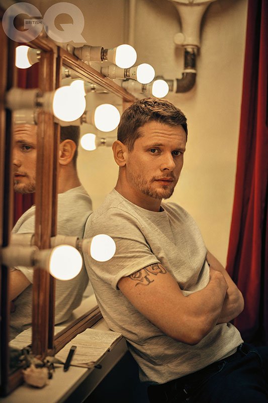 Jack O'Connell leans nonchalantly against a dressing room mirror lined with bright light bulbs. His arms are folded and a tattoo can be glimpsed on his bicep beneath his tshirt sleeve. A rich red velvet curtain hangs behind him. The 'British GQ' logo is embedded over the top left hand corner.