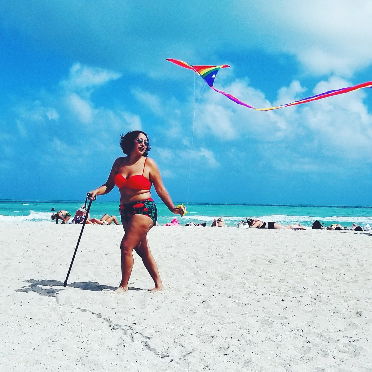 Annie walking with a cane on a sandy beach, wearing a red and black 2 piece swimsuit and flying a rainbow kite with blue skies in the background]