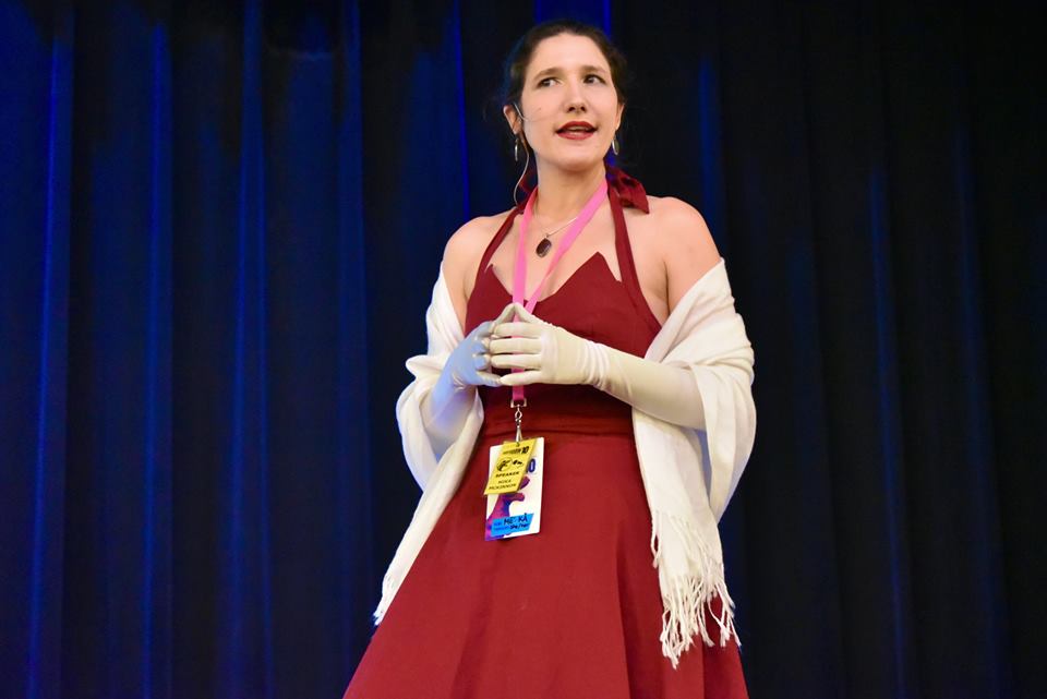 Mika looking only slightly evil while wearing a fancy dress giving a presentation.