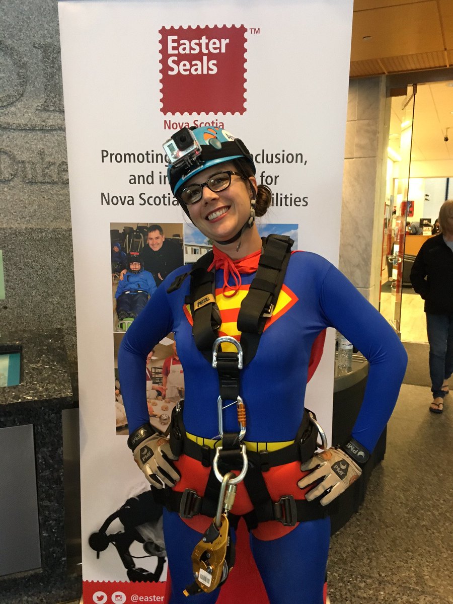 Laura Bain, dressed in rappel gear and a super woman costume, stands in front of a sign for Easter Seals Nova Scotia.