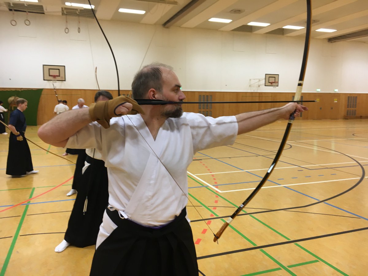 A bearded person with an asymmetric long bow, in full draw. The right hand has a glove. The arrow is pressed to the face. The clothing is japanese Hakama and GI.