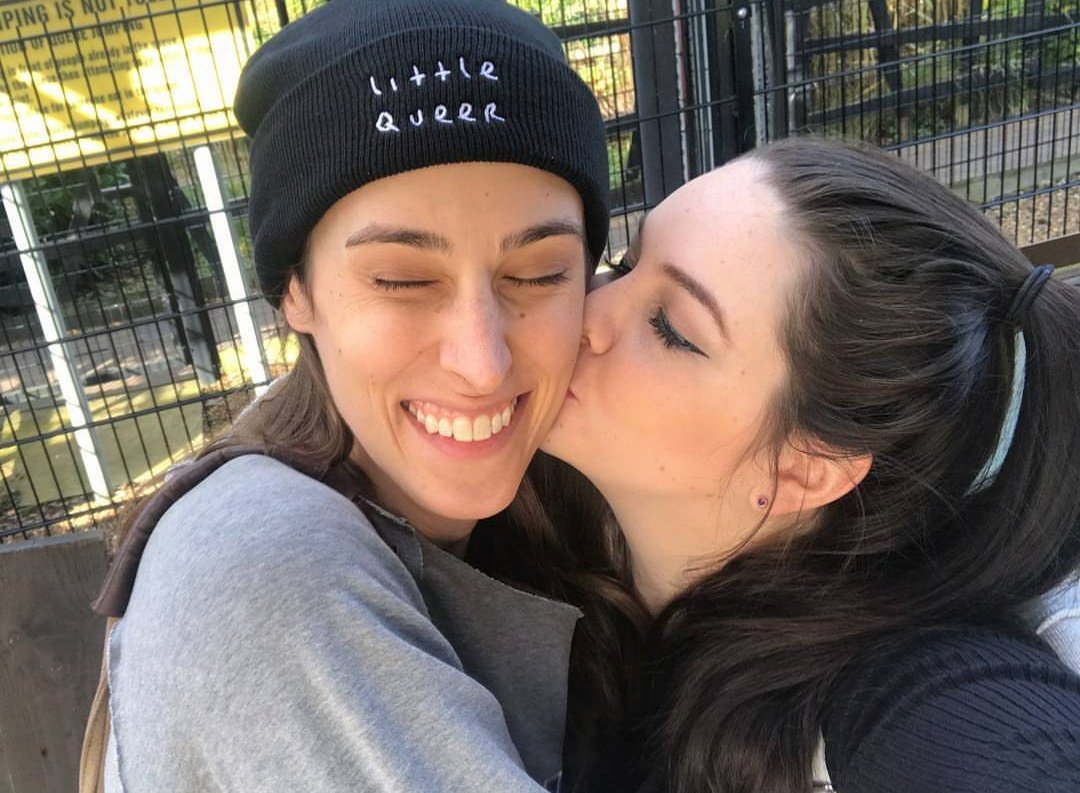 Fiona kissing Riley on the cheek while Riley wears a beanie that says "little queer"