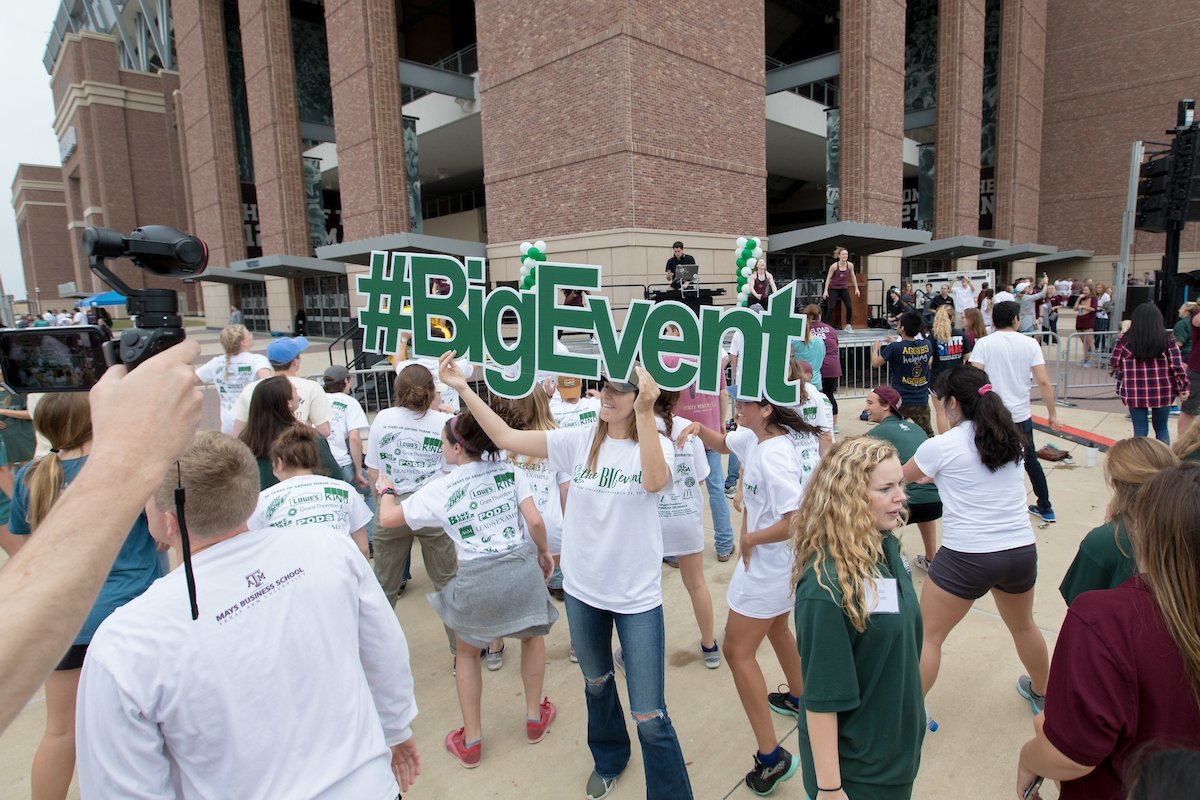Girl standing with a green sign reading: #Big Event surrounded by volunteers