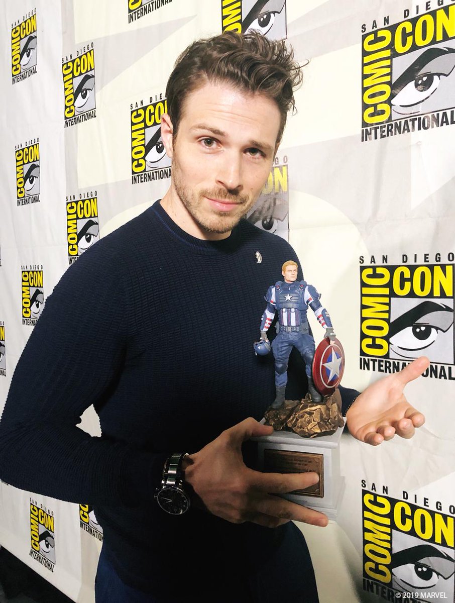 A photo of Captain America performance actor Jeff Schine holding a statue. The statue features Captain America as seen in Marvel's Avengers, posing on top of a pedestal, holding his helmet in one hand, and shield in the other, looking off into the distance.