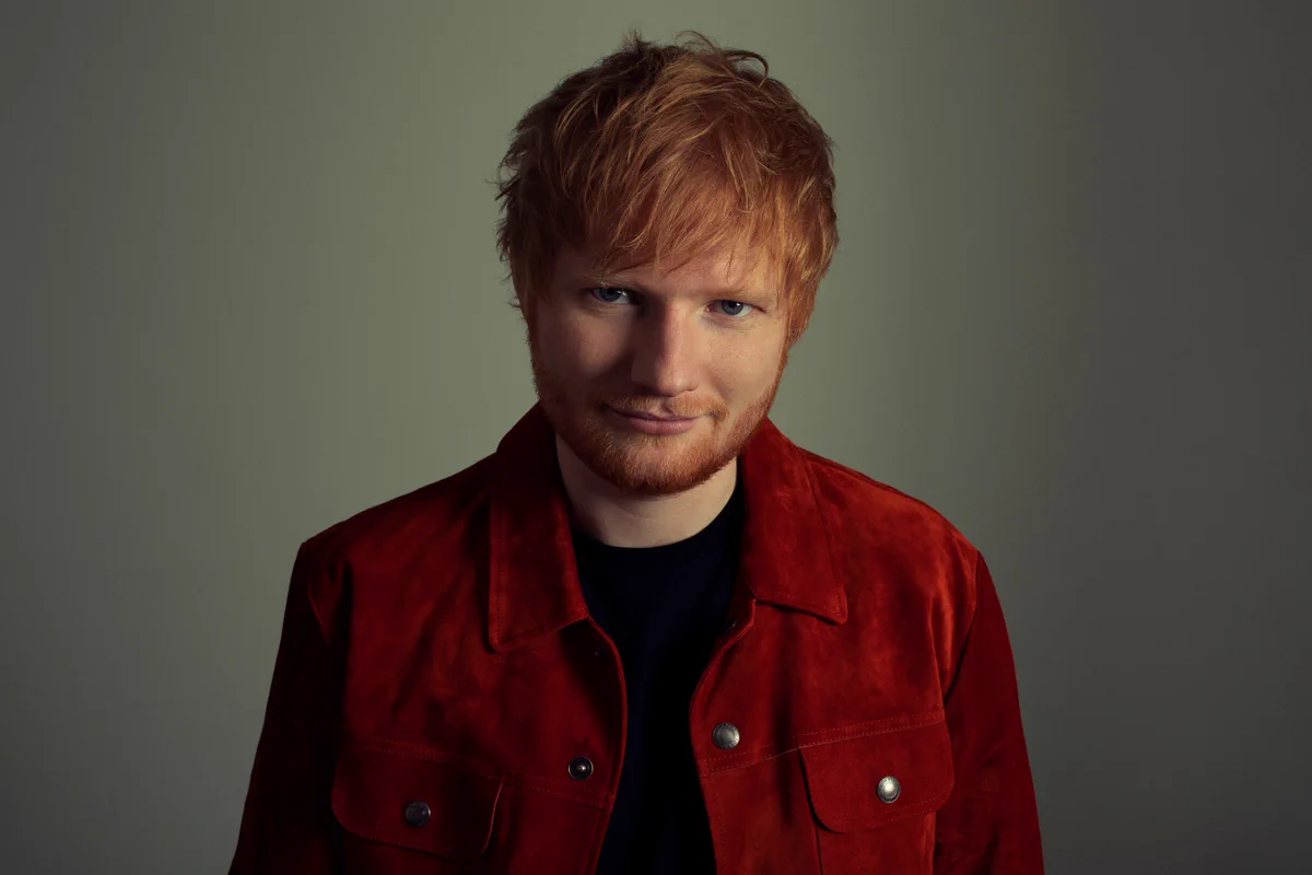 Photo of Ed Sheeran wearing red jacket against green/grey background