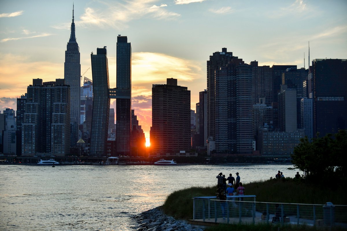 The sun sets between the buildings of the Midtown Manhattan skyline. A crowd across the waterway looks on and takes photos of the view from Hunter's Point South Park.