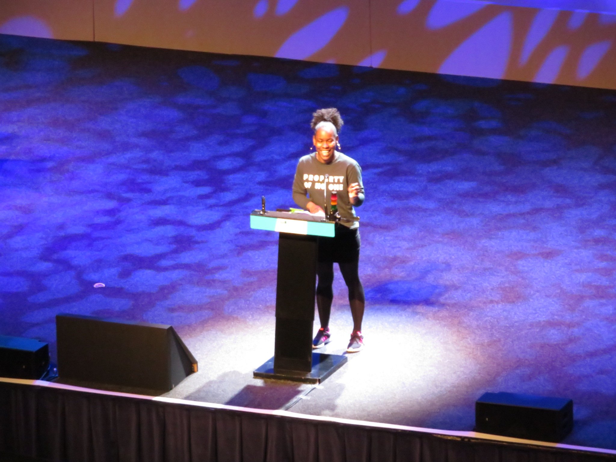 President of NUS in black jeans and grey sweatshirts stands at a lectern on a blue lit stage