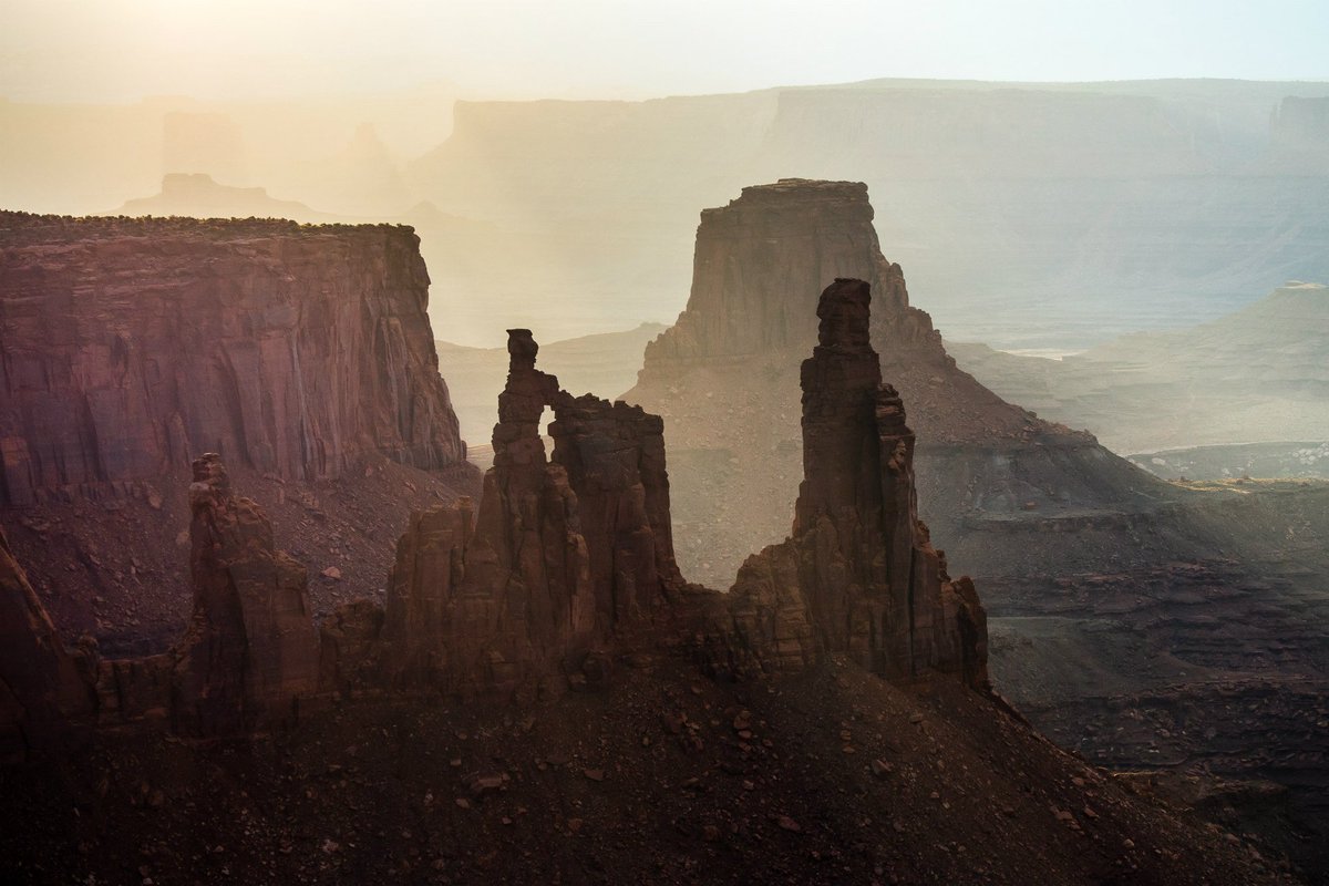 Bright sunbeams shine down through haze onto rocky cliffs and thin stone towers in a wide desert canyon.