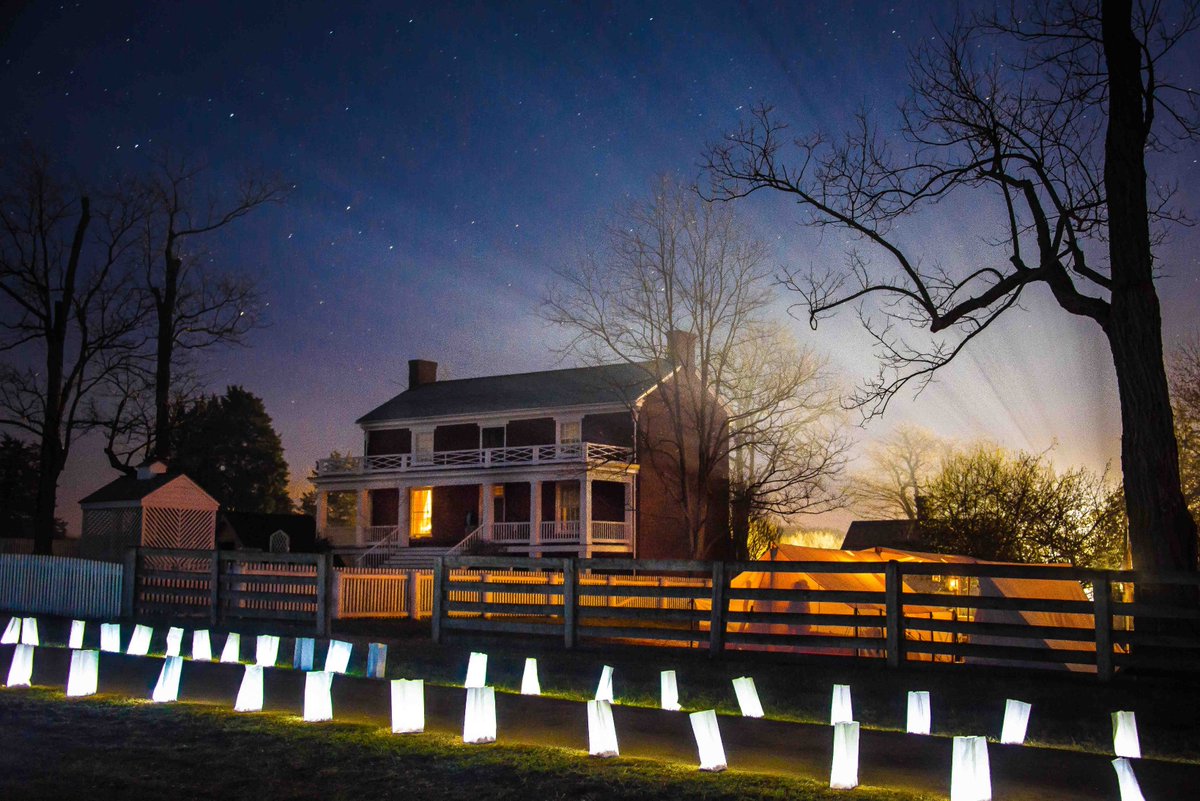 Soft light glows from candles and a campsite next to a large brick home surrounded by a wooden fence under a starry sky.