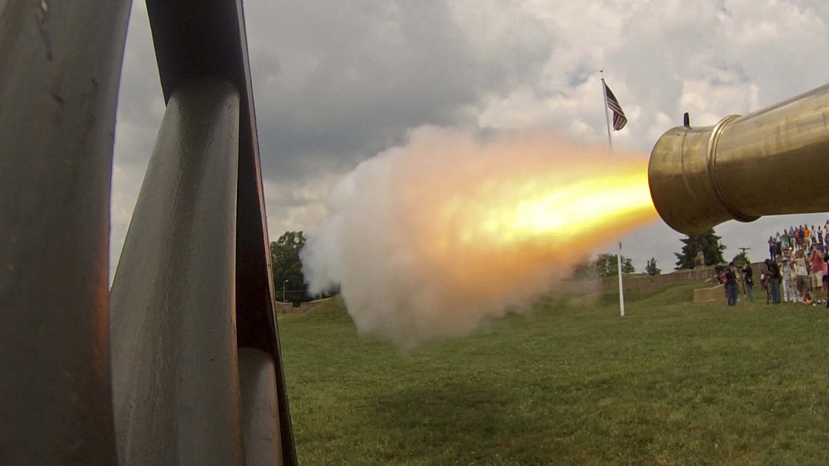 Photo of fire and smoke coming out of the barrel of a canon being fired at a demonstration on a grassy field.