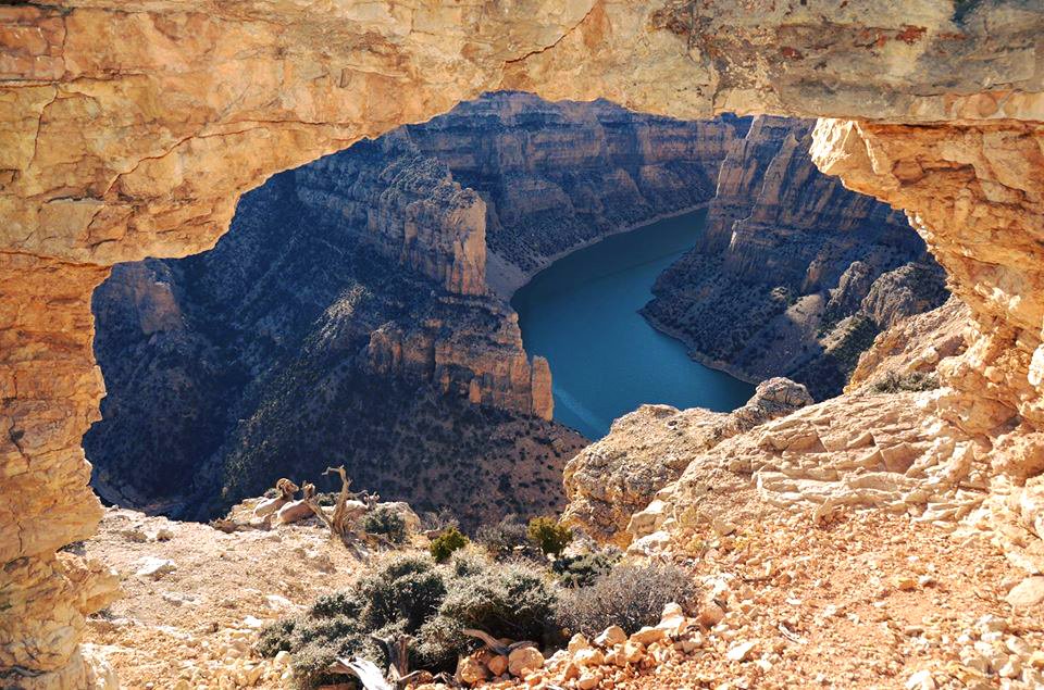 Looking through a natural stone arch, the view shows a narrow, curving canyon with steep walls and a calm, blue river flowing at the bottom.