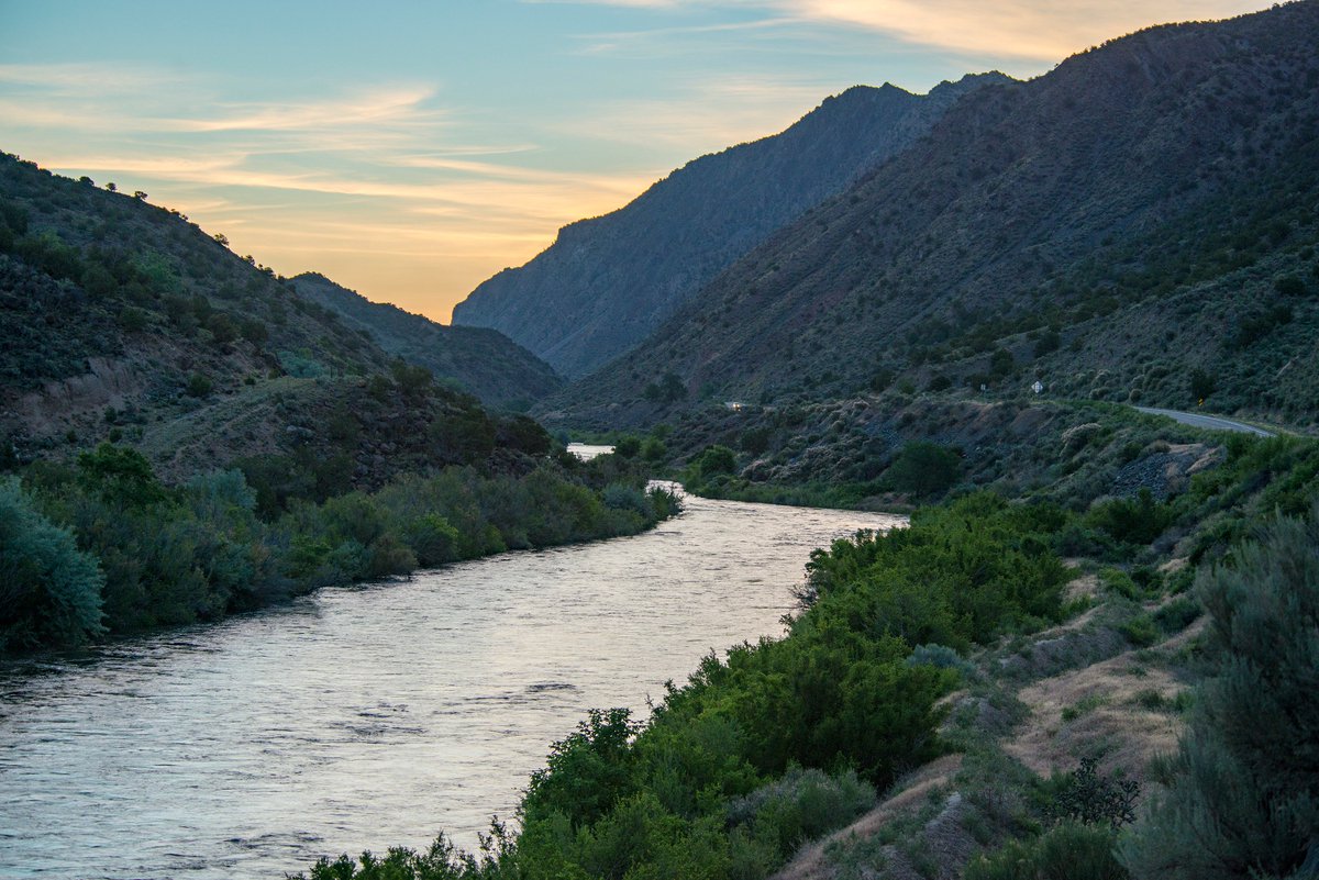 The Rio Grande winds through gentle valley hills on both sides as the soft light of sunrise hits the wispy clouds above.