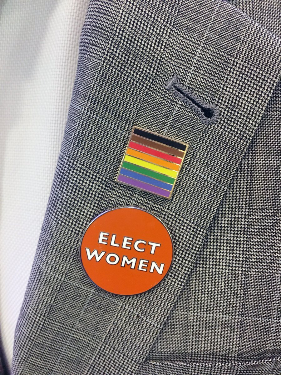 A blazer with two enamel pins. The top pin is a rainbow (inclusive of black and brown stripes) and the bottom is an orange circle that says "ELECT WOMEN."