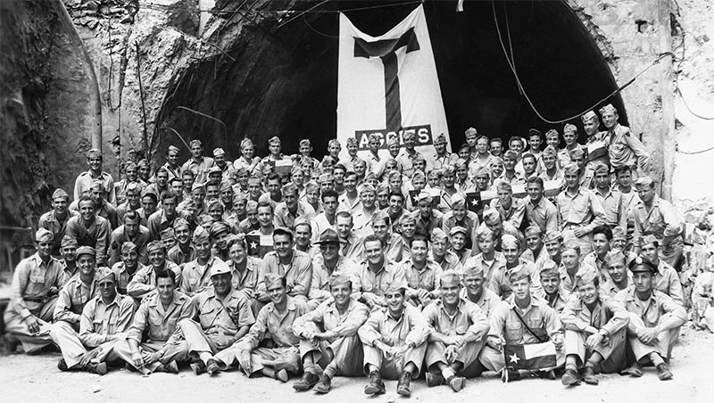 Aggie soldiers sitting on the ground in front of an Aggie flag hanging over a tunnel on the island of Corregidor during world war two