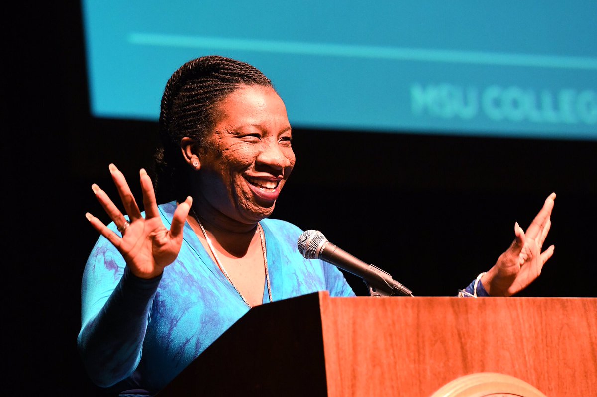 Women in blue dress with hands out speaking into a microphone on stage