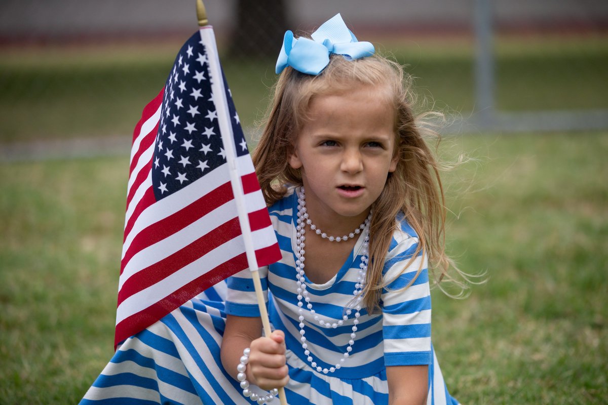 little girl wearing a blue dress and pearls while holding an American flag