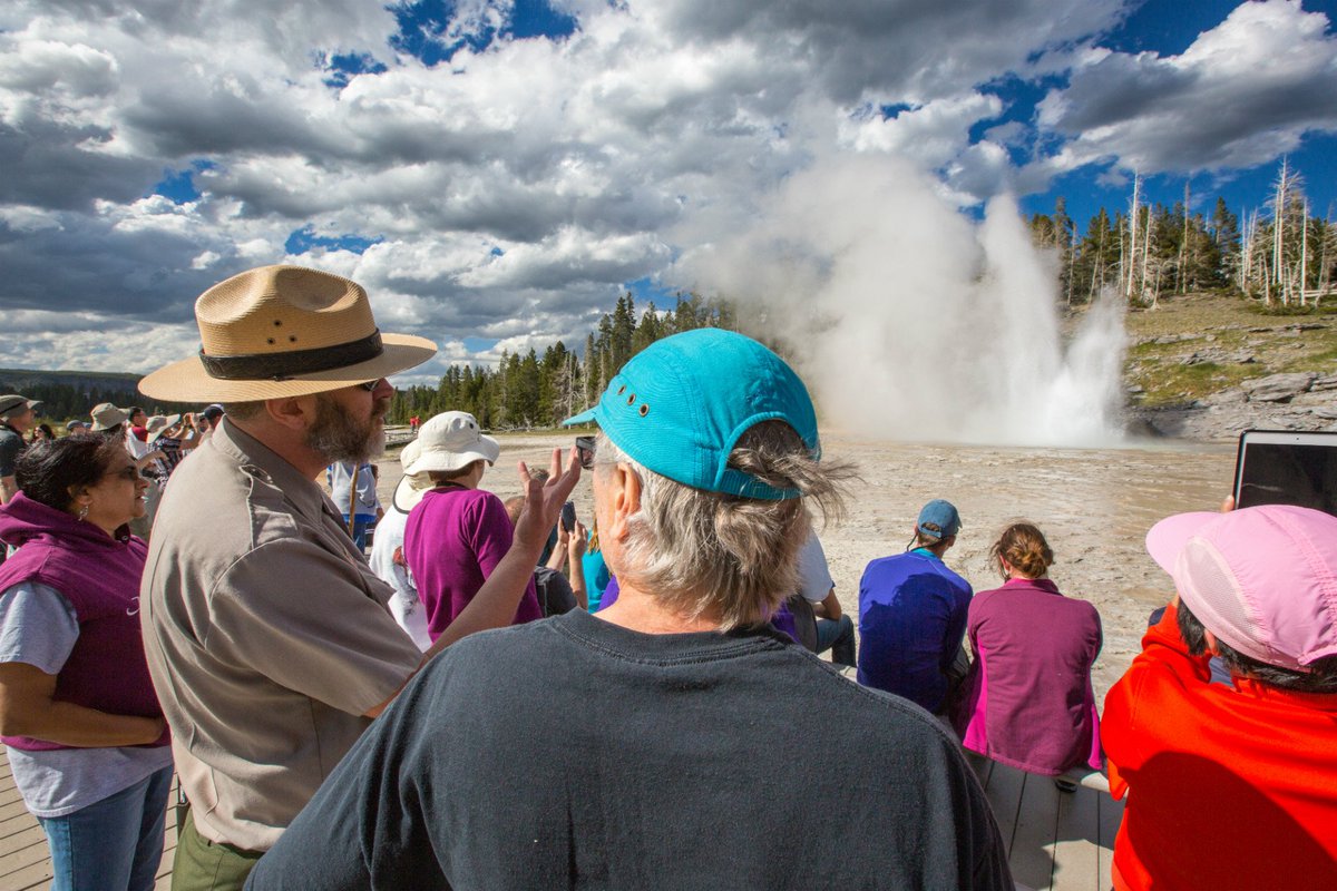 A national park service ranger in a gray uniform with a big hat talks to a group of people watching a geyser erupt in a cloud of steam.