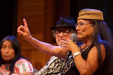 A Native American woman speaking with a microphone with her hand in the air.