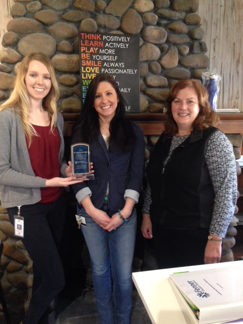 Staff from Fenner Drives, OVR and the Next Step Program smile for a photo. An OVR staff member is holding Fenner Drives’ business award. The individuals in the photo are standing in front of a fireplace.
