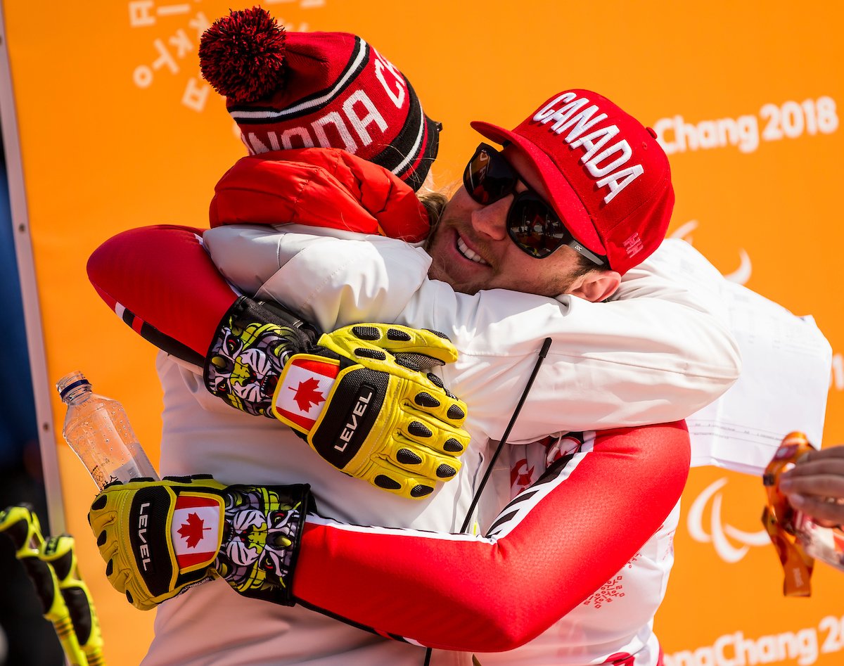 Two athletes wearing Team Canada gear hug in front of a PyeongChang 2018 banner.