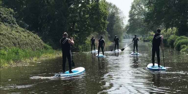 Rear view of the gang stand-up paddle boarding on a tree-lined canal in Chichester.