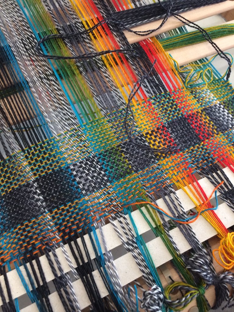 The start of a woven piece of fabric that is still on the loom, it is making squares of different colours with black and white squares.