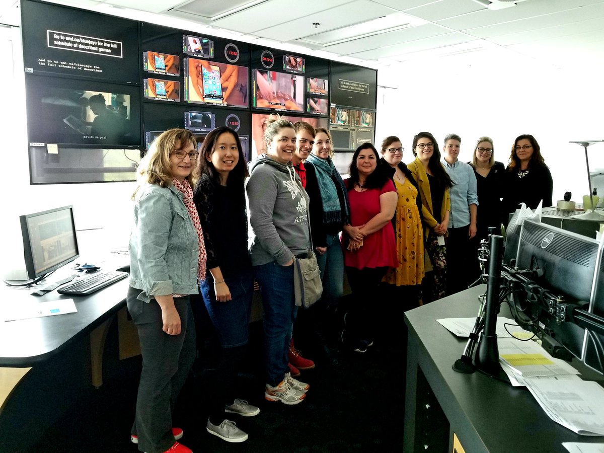 Eleven people stand in a row smiling in front of television screens in AMI Master Control.