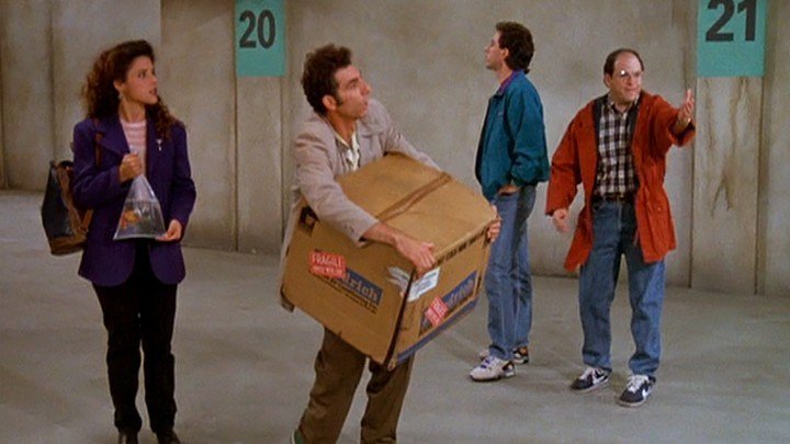 Characters from Seinfeld stand in a parking garage. From left to rights is Elaina Benes holding a gold fish in a bag, Kramer holding a big cardboard box, Jerry Seinfeld looking off into the distance, and George Costanza pointing his hand out.