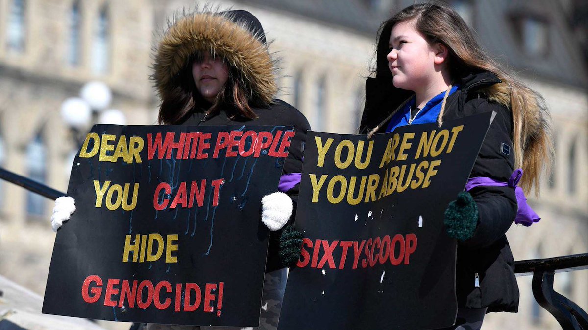 A photograph of two teenage girls holding signs that state: "Dear white people you can't hide genocide" & "You are not your abuse, Sixties Scoop"