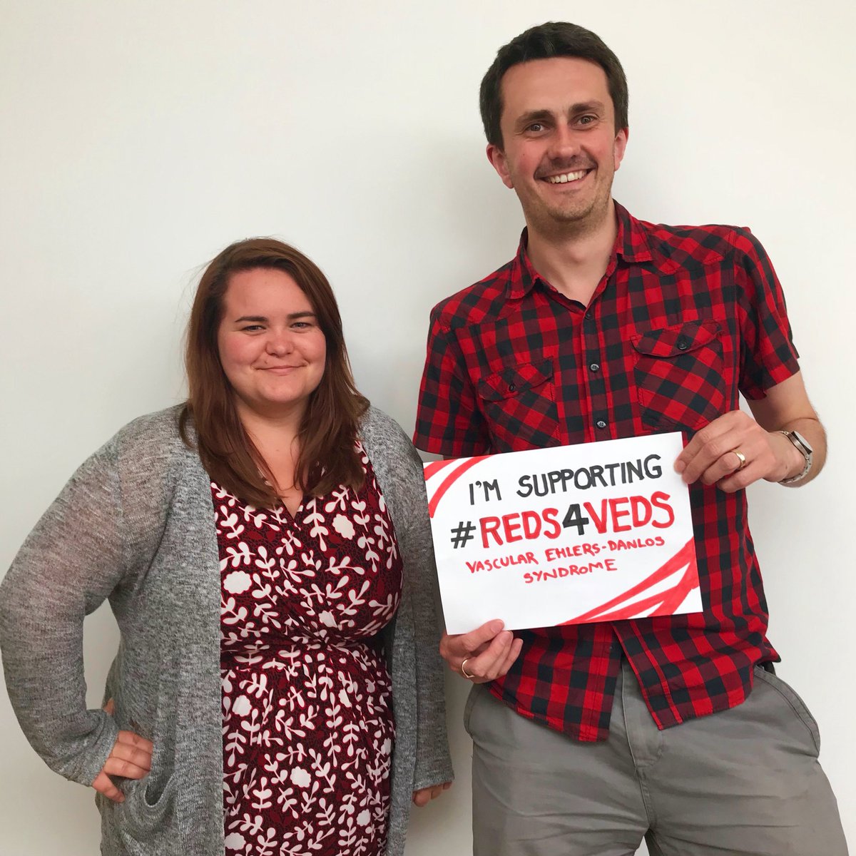 A photo of two people. On the left is a white woman with brown hair and a smile on her face. She wears a red dress with white flowers and a grey cardigan. On the right is a tall white man with brown hair and light stubble. He wears a red and black chequered shirt and grey trousers. He holds a sign that reads “I’m supporting #Reds4Veds” written in red. The man is tall and the woman comes up to his shoulder.