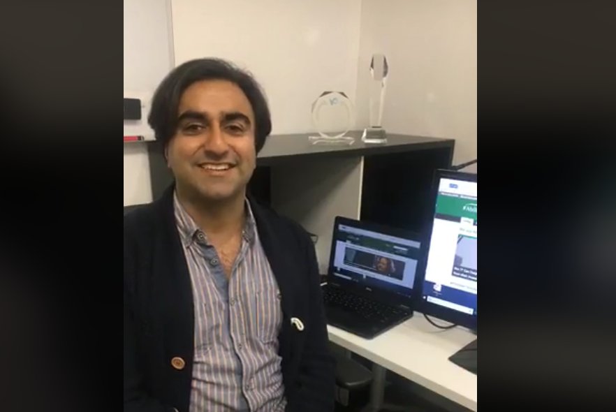 Adi smiling at the camera in an office with computer screens behind him