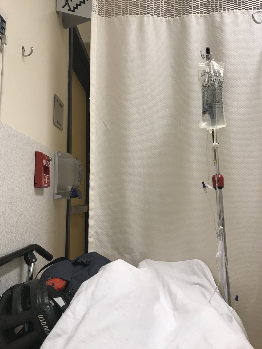 View of the foot of a gurney with a white sheet, a curtain hanging around it, and an IV pole.