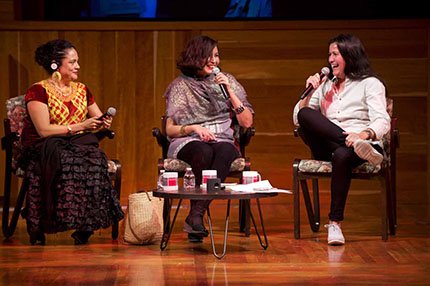 three woman talking on a stage facing each other, holding mics and smiling.