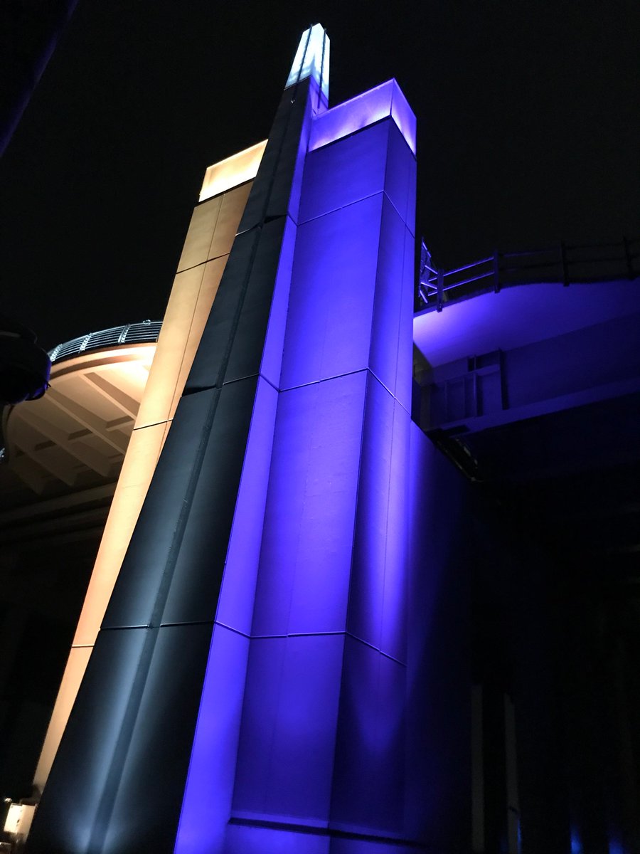 Looking up from the base of a tall concrete tower, half of which is lit purple and half of which is lit gold.